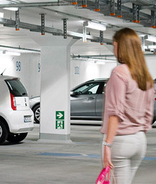 Seorang wanita berjalan menuju mobilnya di garasi parkir hijau yang cerah. - pencahayaan untuk retail