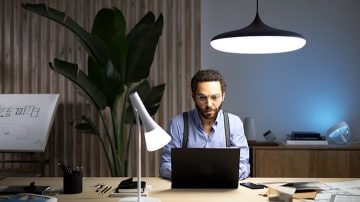 A man sits at a desk with a laptop in front of him, illuminated by a suspended light from above and a table lamp to his right