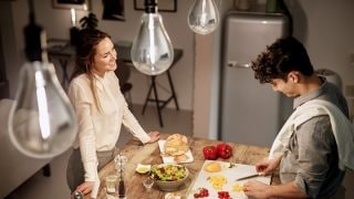 A woman watches a man cutting vegetables on a chopping board in a kitchen, you can see light bulbs in the upper part of the image