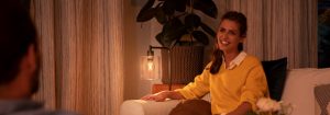 "Suasana ruang keluarga yang nyaman tercipta dari lampu meja LED yang hangat, menyinari wanita bersweater kuning yang tengah menikmati percakapan di malam hari."